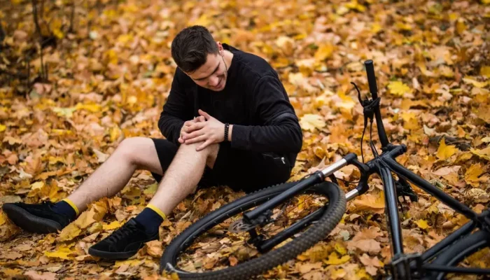 Active young man holding by his hurt or broken leg while lying on autumn forest path by his bicycle